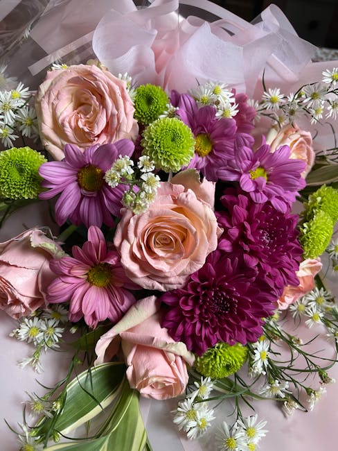 A vibrant bouquet featuring soft pink roses, bright purple daisies, lime green chrysanthemums, and small white daisy-like flowers with yellow centers. The arrangement includes various greenery, such a