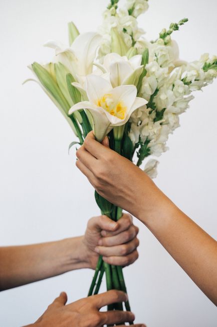 A close-up of two hands exchanging a bouquet of fresh white and cream lilies and delphiniums against a plain white background. The bouquet features large, elegant lily blooms with yellow stamens and l