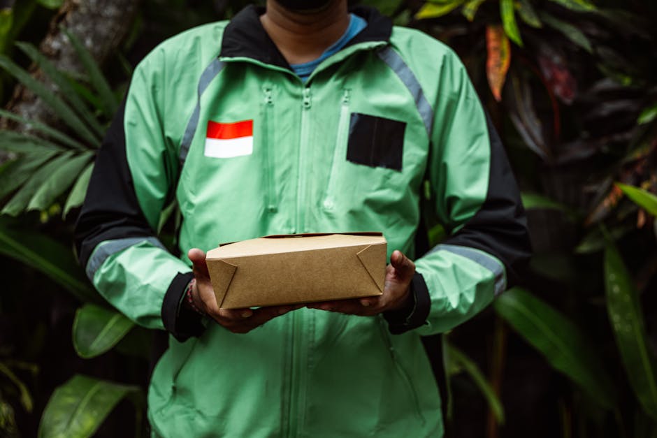 A person wearing a green and black jacket with a red and white flag patch on the chest is holding a rectangular brown cardboard box, likely a flower delivery package, in both hands. The background fea