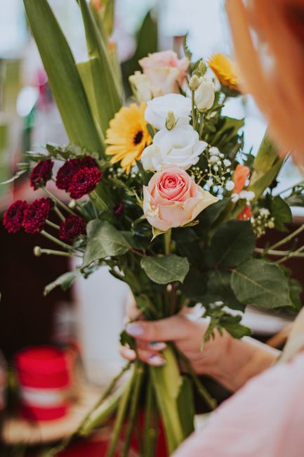 A hand holding a bouquet of fresh flowers arranged with a variety of blooms including pale pink roses, white lisianthus, yellow gerbera daisies, and deep red carnations. The bouquet features lush gree