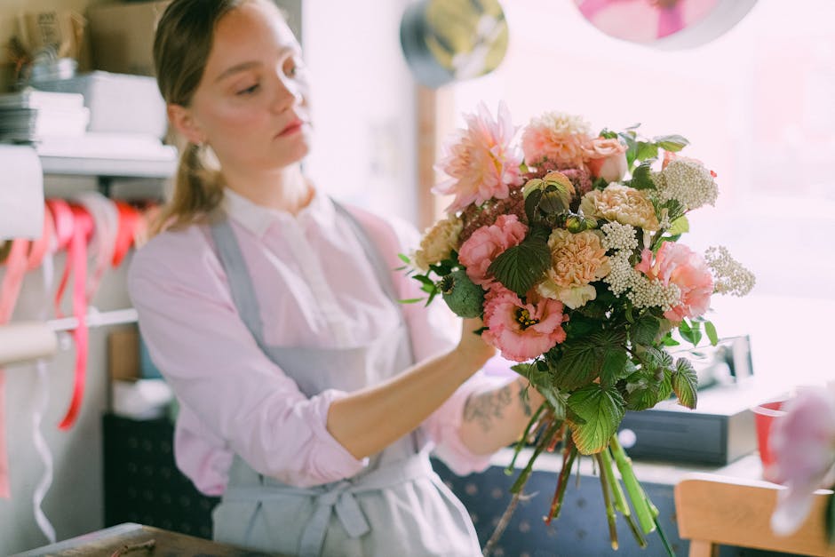 A female florist in a light pink shirt and grey apron carefully arranges a bouquet of fresh flowers in a floral workshop. The bouquet features a mix of pink and cream blooms, including dahlias, carnat
