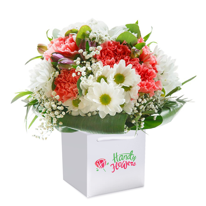 Nestled upright in a pristine, square white gift bag with neat ribbon handles, this expertly arranged bouquet brings together layers of colour and celebration-capturing the essence of special moments shared in Bellingham. The composition is anchored by plush coral and soft pink-red carnations, each petal curling delicately and contributing to a feeling of lush abundance. Between these, clusters of pure white daisy-like chrysanthemums shimmer, their lime-green eyes catching the light like the first blooms of spring in Lewisham's local gardens. Sprigs of white baby's breath wend their way through the arrangement, imparting an airy softness and enhancing the sense of gentle movement, as if the bouquet has just been gathered from a morning market stroll. Ringing the foundation, broad, glossy leaves form a natural collar that supports the dome-like fullness-each leaf inviting the eye to linger. Scattered within, hints of yet-to-open buds-subtle purples and blush pinks-create a layered effect of depth and anticipation. The immaculate gift bag stands out in sharp relief against a brilliant white background, its functional elegance reminiscent of the careful presentation that defines Bellingham's most trusted florists. This lush offering is ready to brighten any celebration-a birthday party along Bromley Road, a new arrival in the neighbourhood, or a heartening 'just because' gesture. Every detail, from flower choice to structure and finish, reflects the craftsmanship and pride of Florist Bellingham, making every delivery feel bespoke and full of meaning.